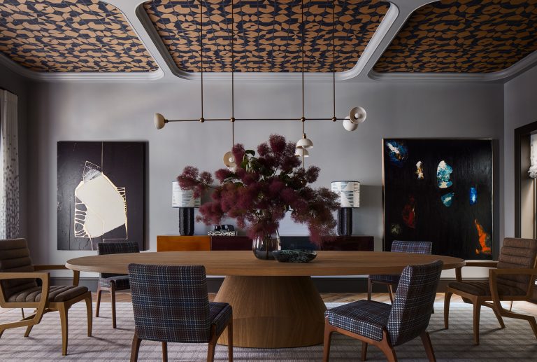 A modern dining room with a round wooden table surrounded by plaid-upholstered chairs. A large vase with dark purple florals sits at the center. Abstract art hangs on gray walls, geometric ceiling patterns, and mid-century lighting complete the stylish look.