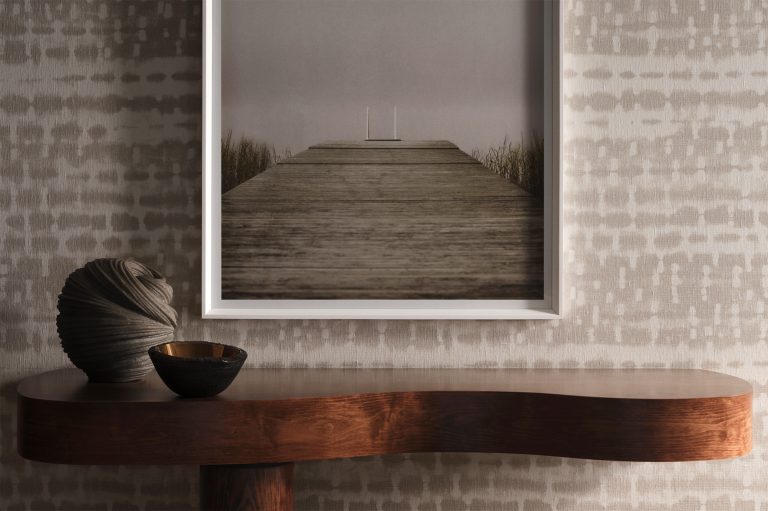 A framed photo of a wooden dock over water hangs above a wavy wooden console table. On the table are a sculptural vase and a small metallic bowl, set against a light wall with a subtle, abstract dotted pattern.