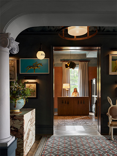 A warmly lit interior shows a reception desk with a person seated behind it, framed by a doorway. The foreground has patterned tile flooring, plants, and eclectic wall art. Vintage decor, large windows, and soft lighting create an inviting, stylish atmosphere.