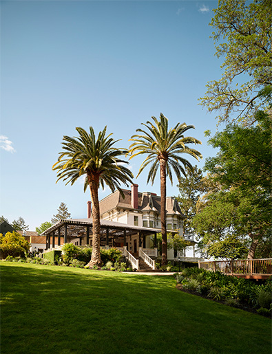 A large Victorian-style house with a porch sits behind two tall palm trees. The building has a steep roof, multiple chimneys, and is surrounded by lush green lawn and trees under a clear blue sky.