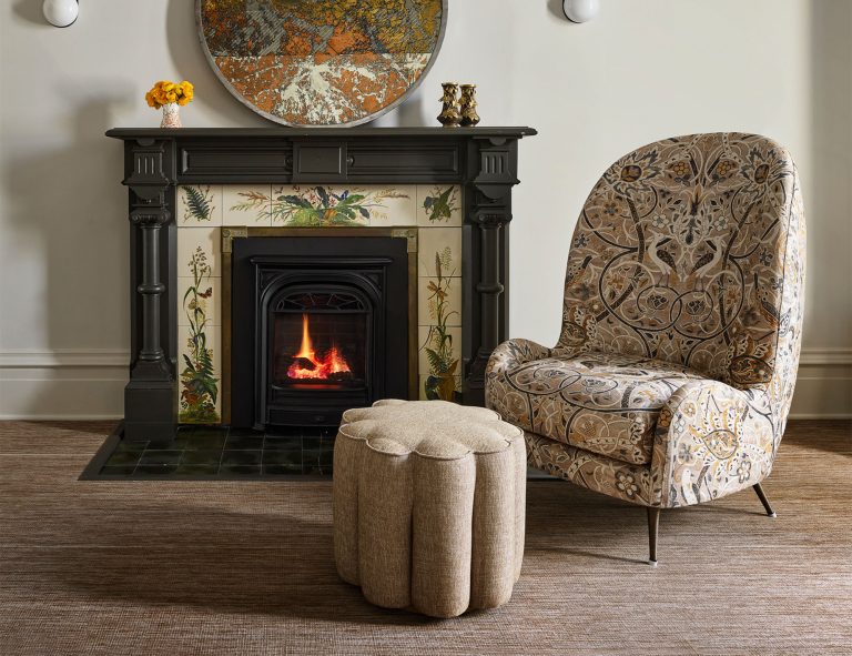 A cozy living room with a black fireplace featuring decorative tiled sides, a small fire burning inside, a round ottoman, and a patterned armchair. Above the mantel is a round, artistic wall piece and two small vases, one with yellow flowers.