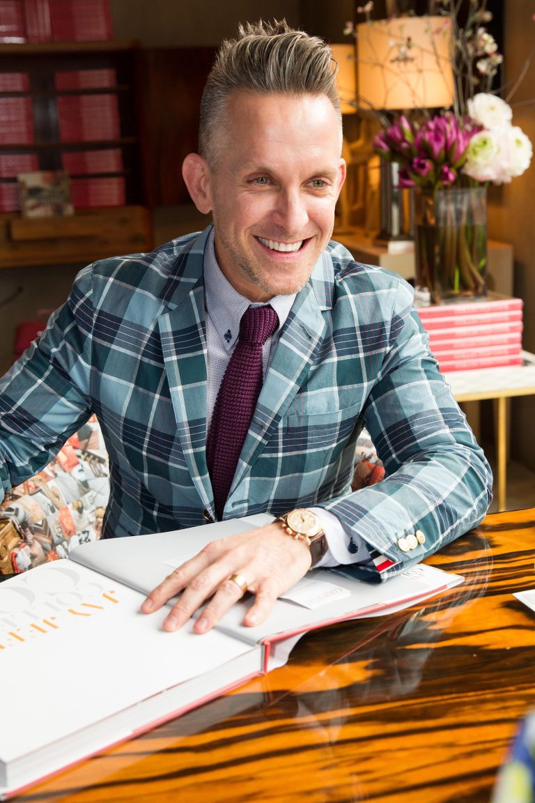 A smiling man with short, styled hair wears a blue and green plaid suit jacket, lavender shirt, and maroon tie. He sits at a polished wooden table with an open book, surrounded by pink books and flowers, in a warmly lit, stylish room.