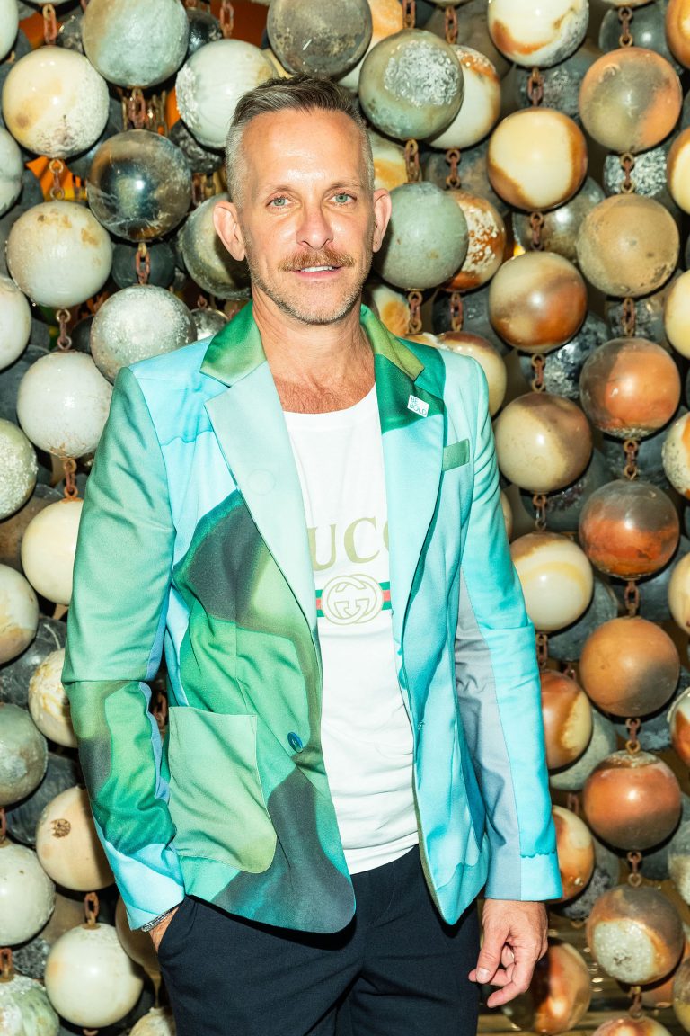 A man with short hair and a trimmed beard wears a teal and green patterned blazer over a white Gucci T-shirt. He stands in front of a background made of round, metallic spheres connected by chains, creating a textured, artistic backdrop.