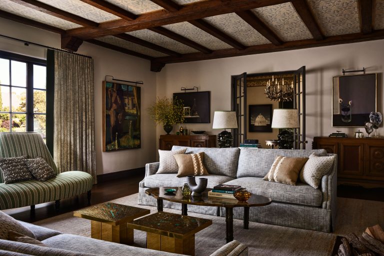 A stylish living room with wood-beamed ceiling, gray sofas with beige pillows, a wooden coffee table with books and decor, armchair by a window with drapes, framed artwork on the walls, sideboard with lamps, and a decorative screen in warm, neutral tones.