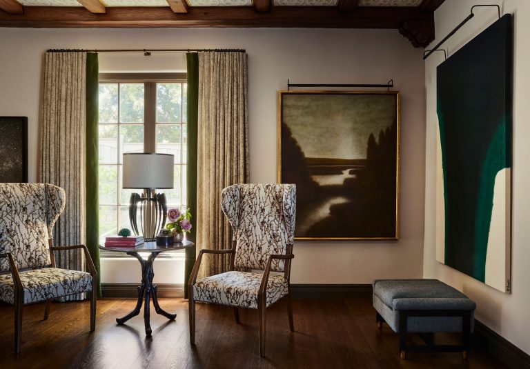 A sunlit living room with two patterned armchairs around a small round table topped by a lamp, books, and a vase. Behind is a window with curtains. On the wall are moody landscape and abstract paintings; a blue ottoman sits below the abstract artwork.