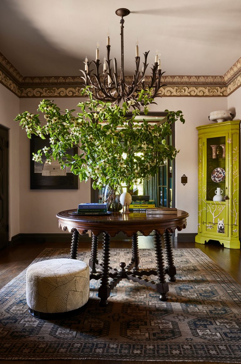 A round wooden table with a scalloped edge and decorative legs stands on a patterned rug. On the table are books, two vases, and a large leafy branch centerpiece. Above hangs a black chandelier; a yellow china cabinet stands against the wall in the background.