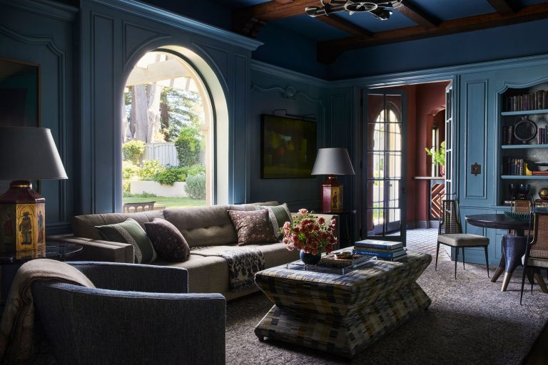 A cozy, elegant living room with blue paneled walls, arched windows and doors letting in sunlight, a beige sofa, patterned coffee table, armchairs, bookshelves, table lamps, and a vase of flowers, creating a warm, inviting atmosphere.