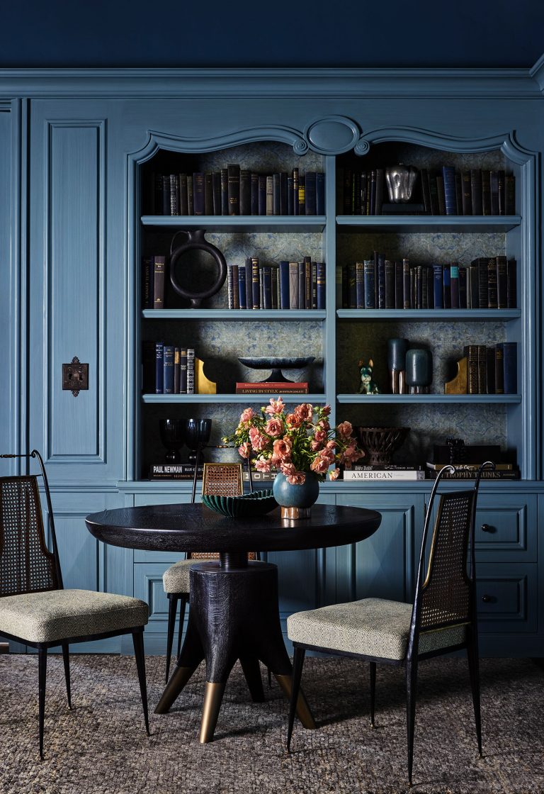 A sophisticated dining area features a round black table with a vase of pink flowers, surrounded by three upholstered chairs with cane backs. Behind, blue built-in bookshelves hold dark books, decorative vases, bowls, and various art objects.