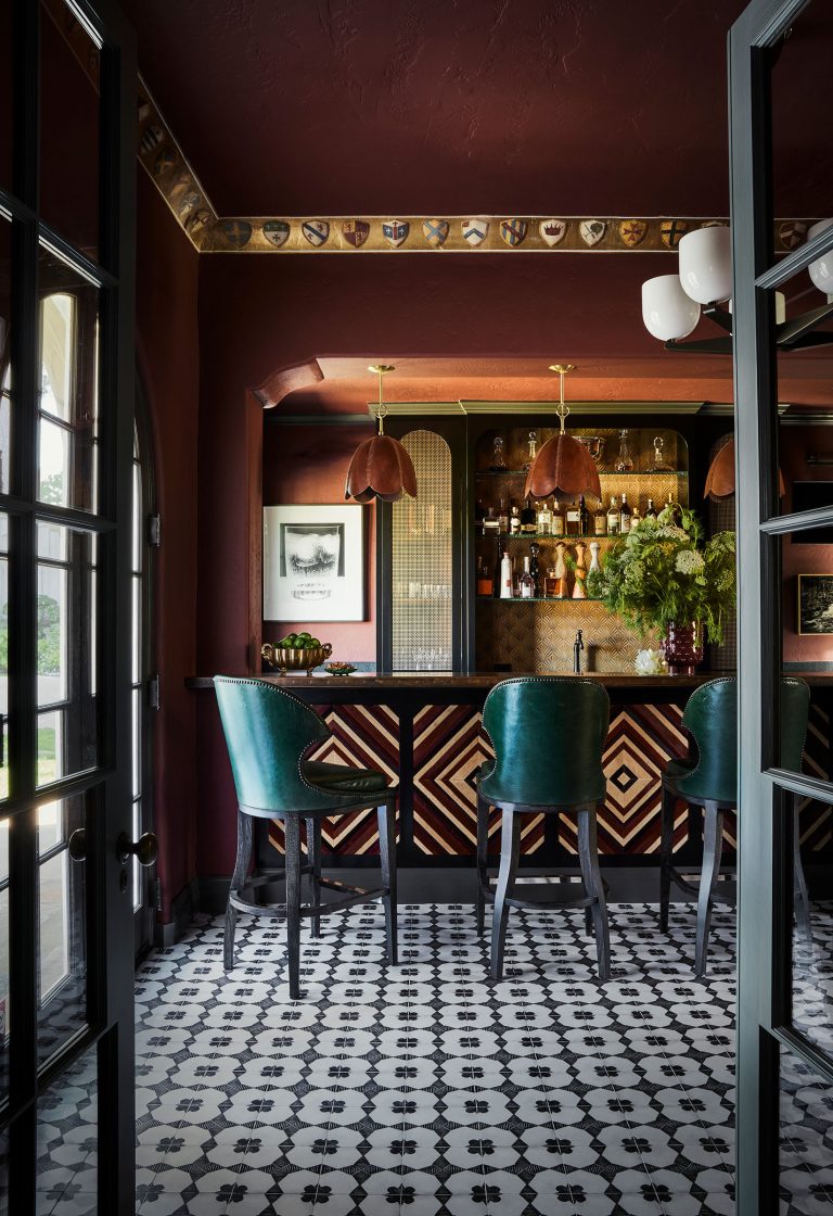 A stylish home bar with geometric wood paneling, green leather barstools, patterned black-and-white floor tiles, and a mirrored back wall with shelves of bottles and glassware. Two pendant lights with copper shades hang above the bar. The room has dark red walls and ceiling.