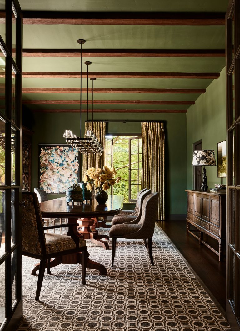 A dining room with green walls and ceiling, wooden beams, a large window with gold curtains, and an elegant wooden table surrounded by patterned chairs. A geometric rug covers the floor, and the room features floral décor and a dark sideboard with a lamp.