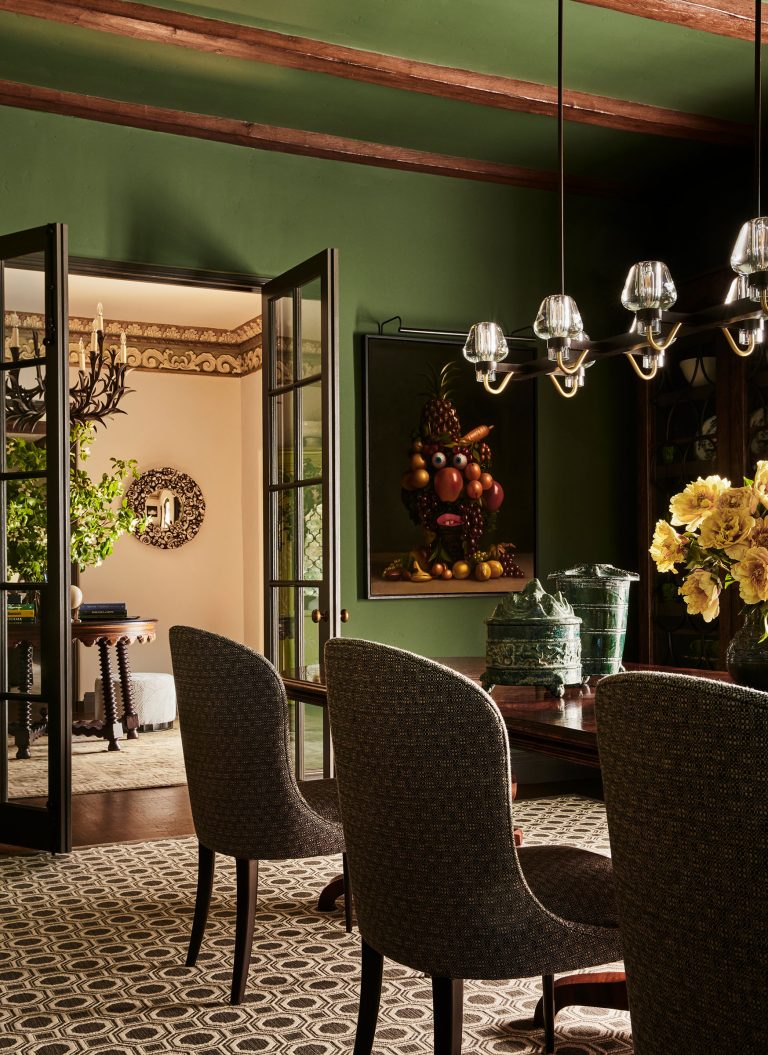 A sophisticated dining room with green walls and wood-beamed ceiling. Four textured chairs surround a dark table topped with yellow flowers. A modern glass chandelier hangs above. In the background, glass doors open to a bright room with a large mirror and leafy plant.