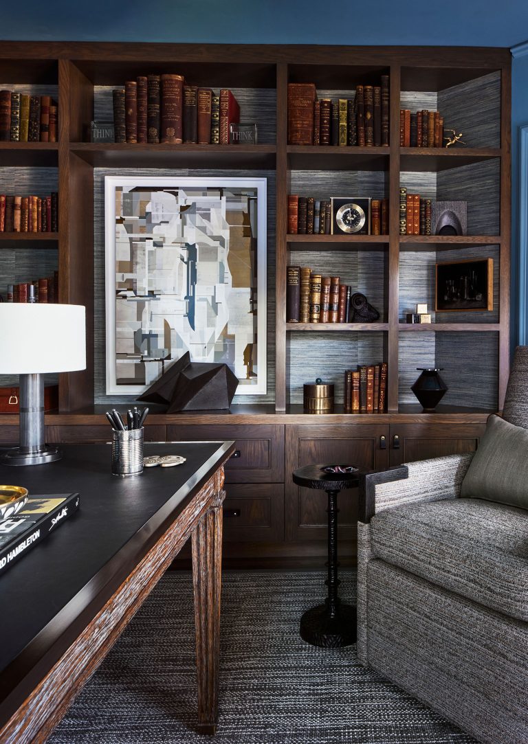 A cozy, elegant library with dark wood shelves filled with old books and art objects, a framed abstract artwork at the center, a plush gray armchair, and a desk with a lamp, pens, and books, all set on a textured gray rug.
