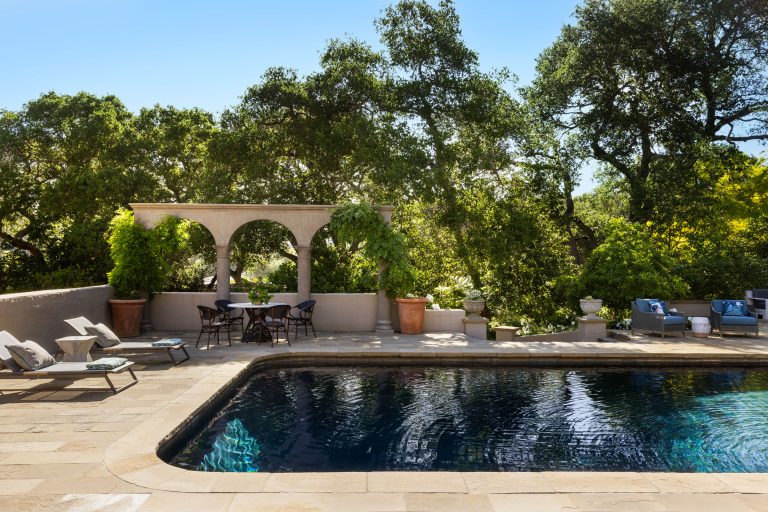 A serene outdoor patio features a rectangular pool surrounded by stone tiles, lounge chairs with cushions, and a shaded dining area under an arched pergola with lush greenery and potted plants, set against a backdrop of leafy trees under a clear sky.