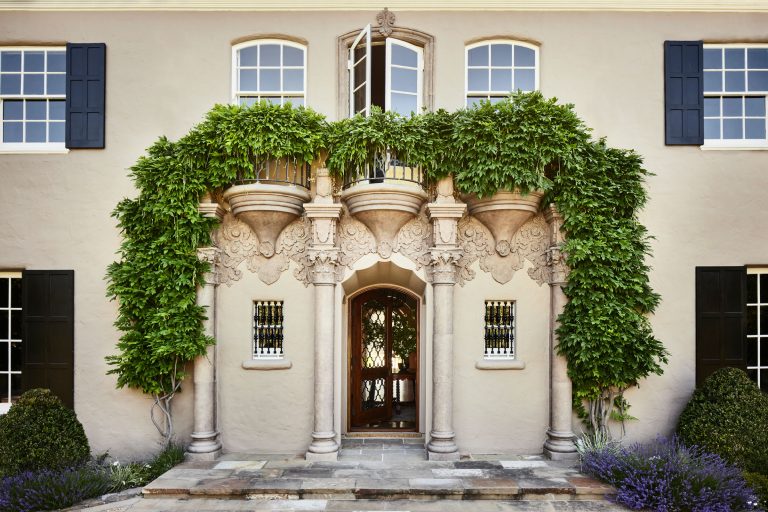 Elegant arched doorway with ornate columns and balcony, framed by lush green vines, on a beige stucco building. Black shutters, manicured shrubs, and lavender plants enhance the symmetrical, classical facade. Upper windows are open above the entrance.