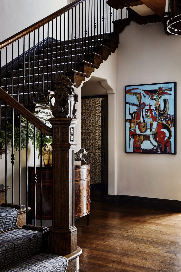 A wooden staircase with dark railings and patterned carpet rises above a polished wood floor. A detailed lion sculpture sits on the newel post. An abstract painting hangs on a white wall. A leafy plant and antique sideboard are tucked beneath the stairs.