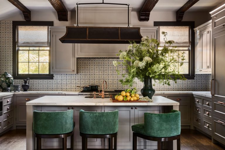 A stylish kitchen with gray cabinets, a white marble island, and three green velvet barstools. A large vase of greenery and lemons sits on the island. Patterned backsplash, wooden beams, and two windows add warmth and light to the space.
