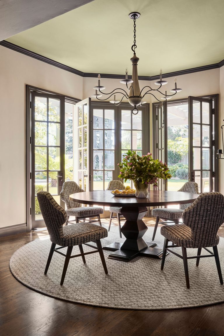 A bright dining room with a round wooden table, six woven chairs, a large floral centerpiece, and a bowl of lemons. French doors open to a sunny outdoor space. Dark wood floors are covered by a round rug, and a chandelier hangs from the ceiling.