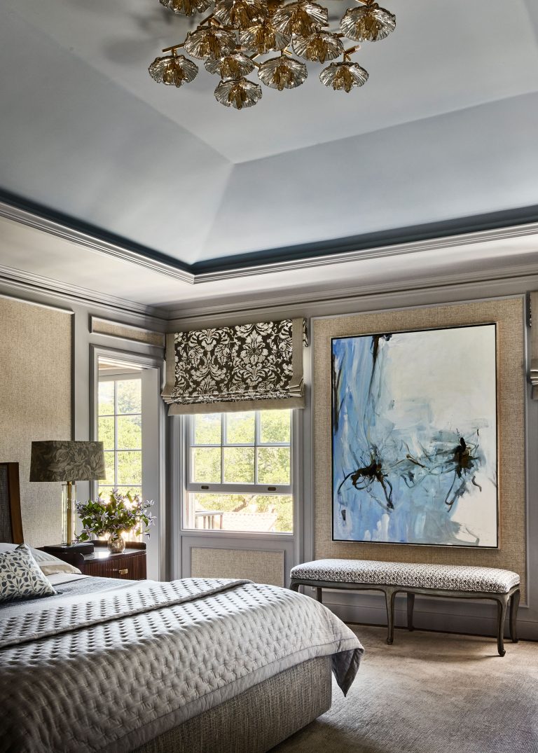 Elegant bedroom with a tufted bed, patterned bedding, and a wooden nightstand holding a lamp and flowers. Large abstract painting on the wall, upholstered bench beneath, floral roman shades on windows, and a gold leaf chandelier overhead. Neutral, calming color palette.