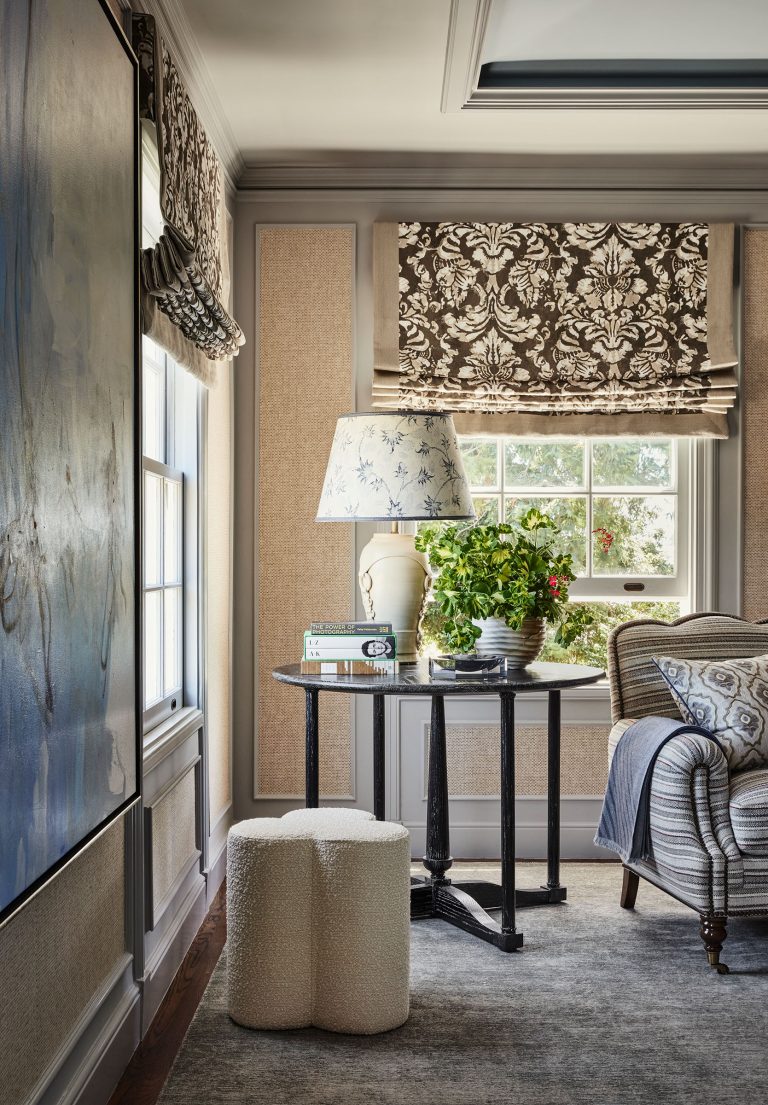 A cozy living room corner features a striped armchair, a black side table with a floral lamp, books, and a potted plant. Roman shades with a damask pattern cover the windows, and textured walls are complemented by large abstract art on the left.