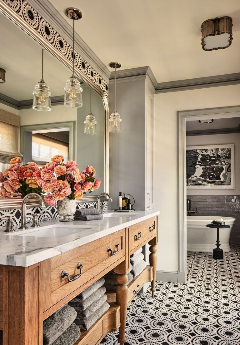 A stylish bathroom with patterned black-and-white tile flooring, a wooden vanity with a marble countertop, towels neatly stacked on shelves, a vase of pink flowers, glass pendant lights, a large ornate mirror, and a view into a tub and art-filled room beyond.