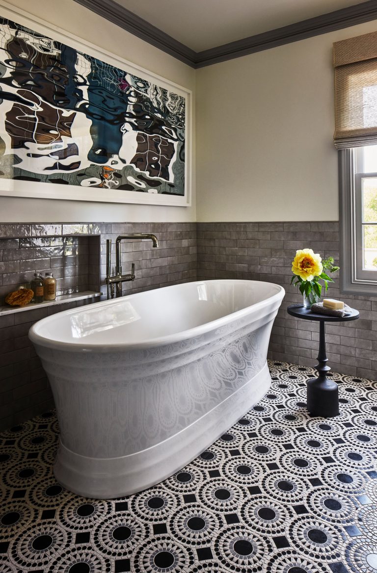 A modern bathroom with a freestanding white bathtub, gray textured wall tiles, and black-and-white patterned floor tiles. Above the tub hangs abstract artwork. A small black side table with a yellow flower in a vase is beside the tub, near a window with a woven shade.