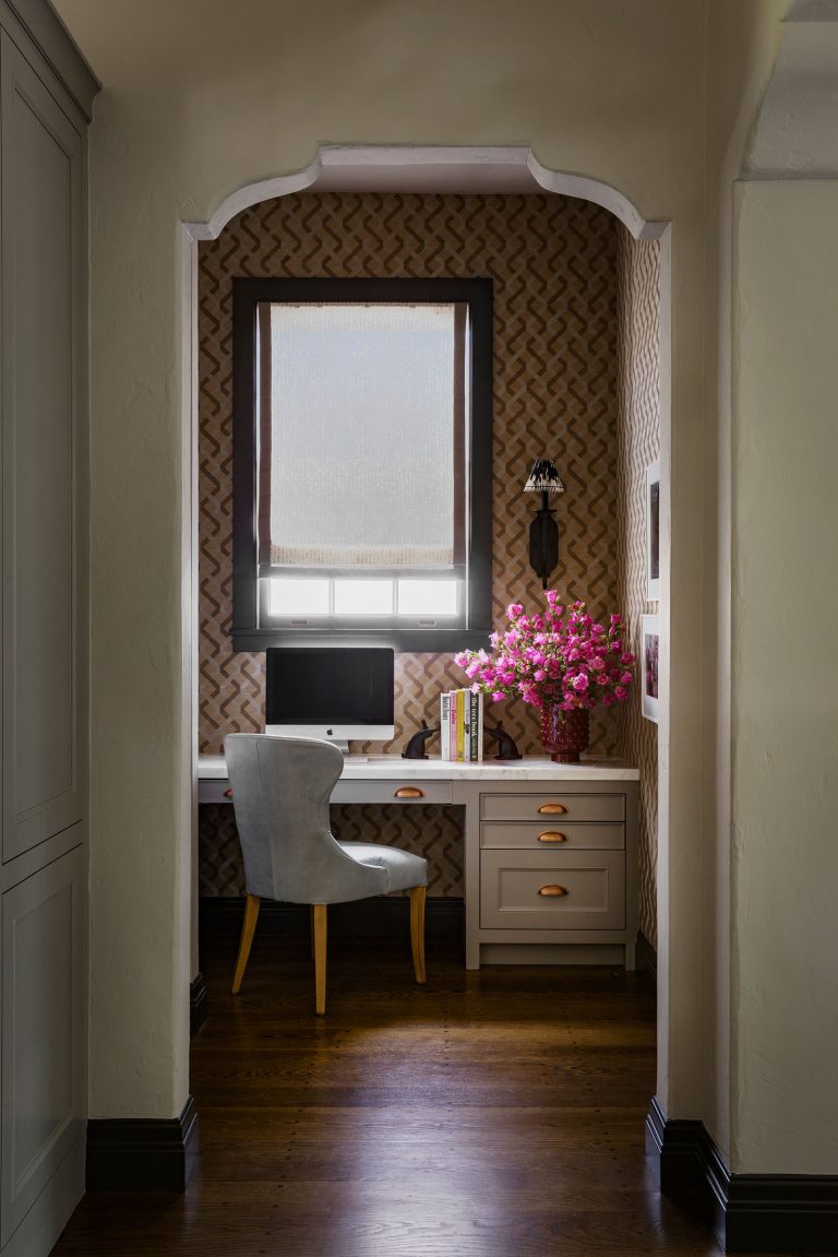 A cozy home office nook with a white desk, gray upholstered chair, desktop computer, a stack of books, and a vase of bright pink flowers. Patterned wallpaper, a window with a shade, dark wood floor, and soft lighting create a warm, inviting atmosphere.