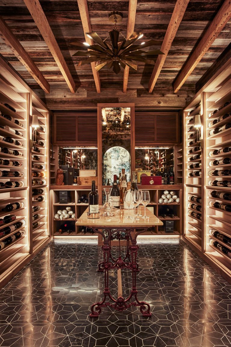 A luxurious wine cellar with wooden racks filled with wine bottles lines both walls. A small table holds wine glasses and bottles. Warm lighting highlights wood beams on the ceiling and a patterned tiled floor. A mirrored wall and archway appear at the back.