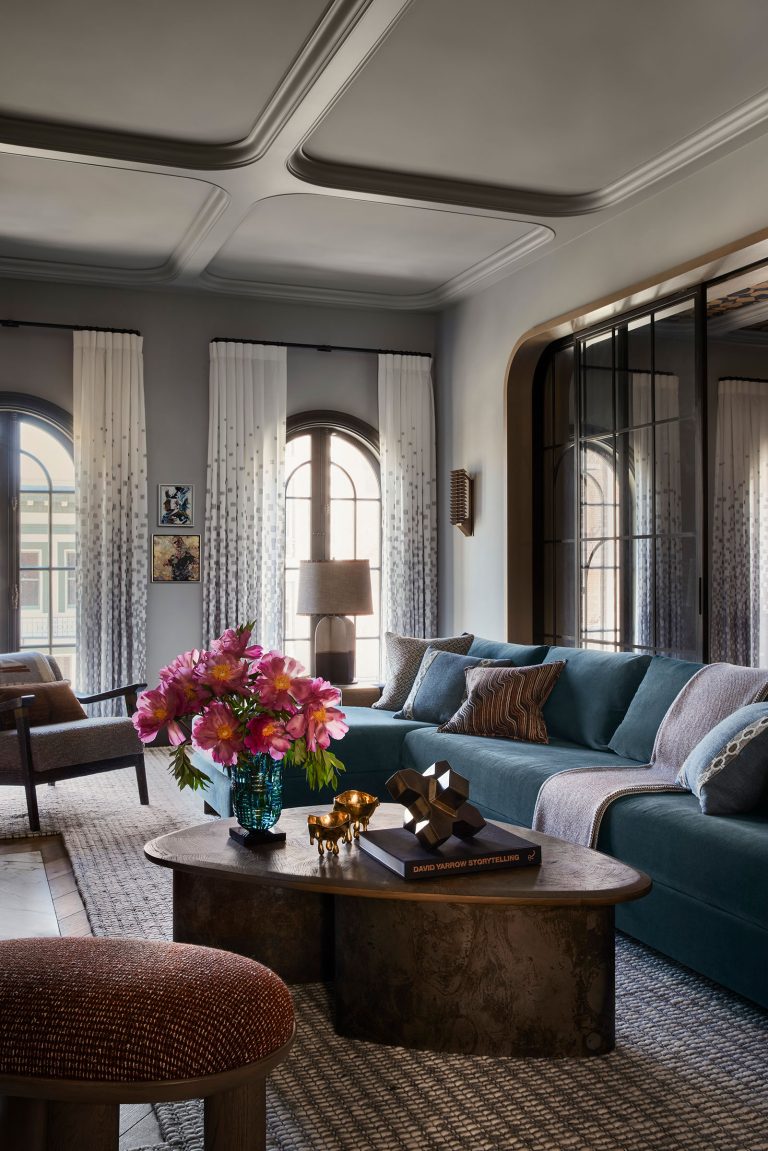 Elegant living room with a teal sectional sofa, patterned cushions, and a round coffee table topped with a vase of pink flowers, decorative objects, and books. Large arched windows with gradient drapes let in natural light, and a textured rug covers the floor.