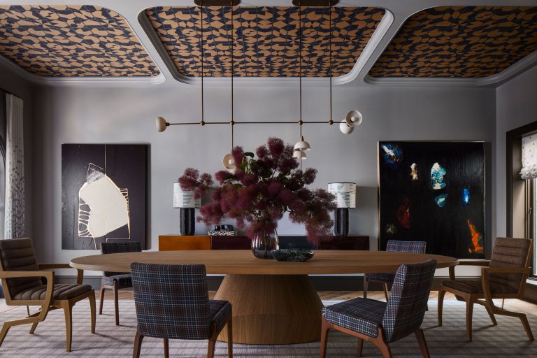 A modern dining room with a wooden oval table, plaid upholstered chairs, and a large centerpiece of leafy branches. The ceiling features patterned panels, and abstract art decorates the gray walls. Two table lamps sit on a sideboard beneath the artwork.