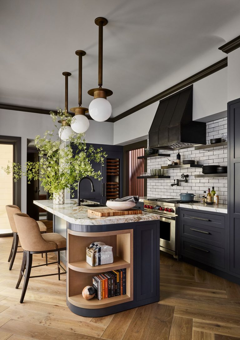 Modern kitchen with a marble island, two beige chairs, and open shelves with books and decor. Three round pendant lights hang above. Dark cabinets, a black range hood, white subway tile backsplash, and wooden accents complete the warm, stylish space. Green branches in a vase.