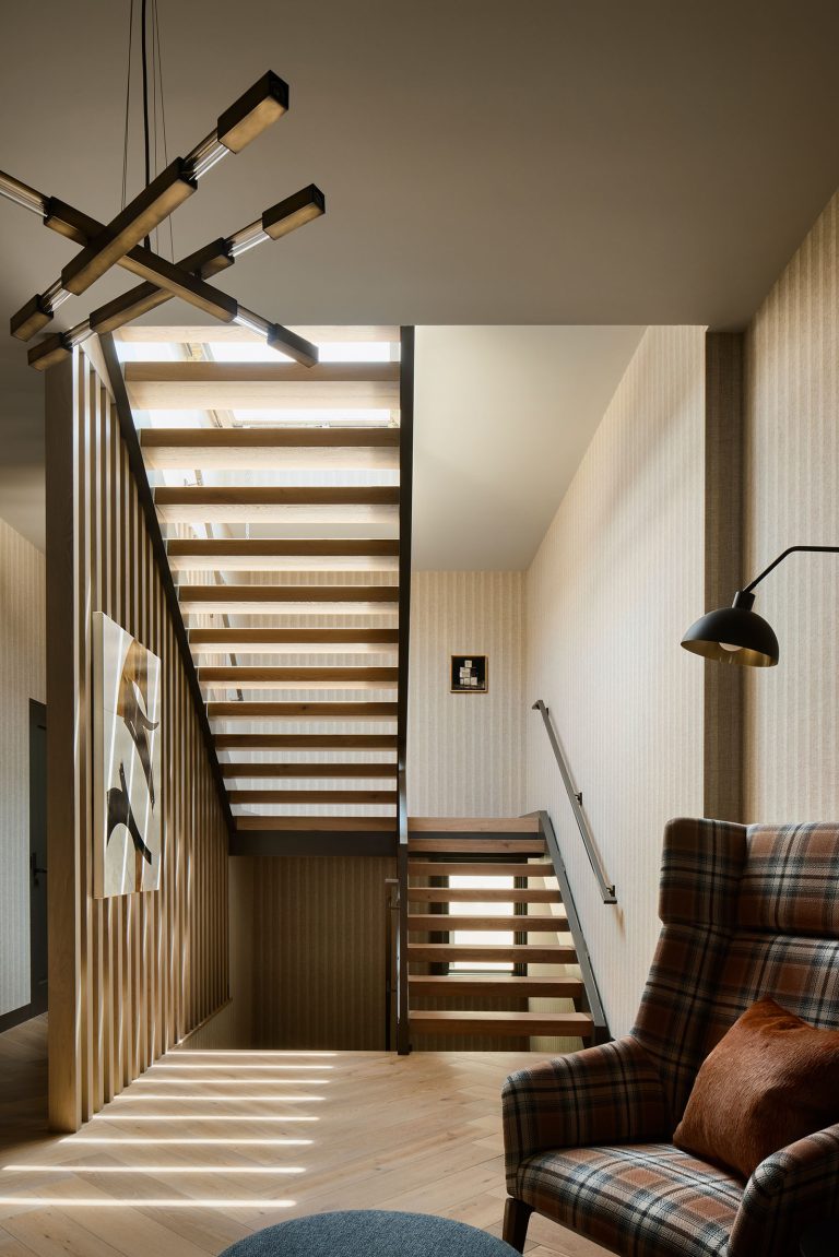 Modern interior with wooden open-riser staircase featuring sunlight casting shadows. A plaid armchair with a brown pillow sits below a black wall lamp. Abstract art hangs on the wall; a geometric pendant light is on the ceiling. Neutral colors dominate the scene.