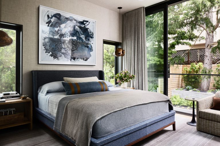 Modern bedroom with large windows, a dark blue upholstered bed, neutral bedding, and abstract art above the headboard. Light pours in, revealing trees outside. A glass vase with flowers sits on the nightstand, and a cozy chair and side table are by the window.