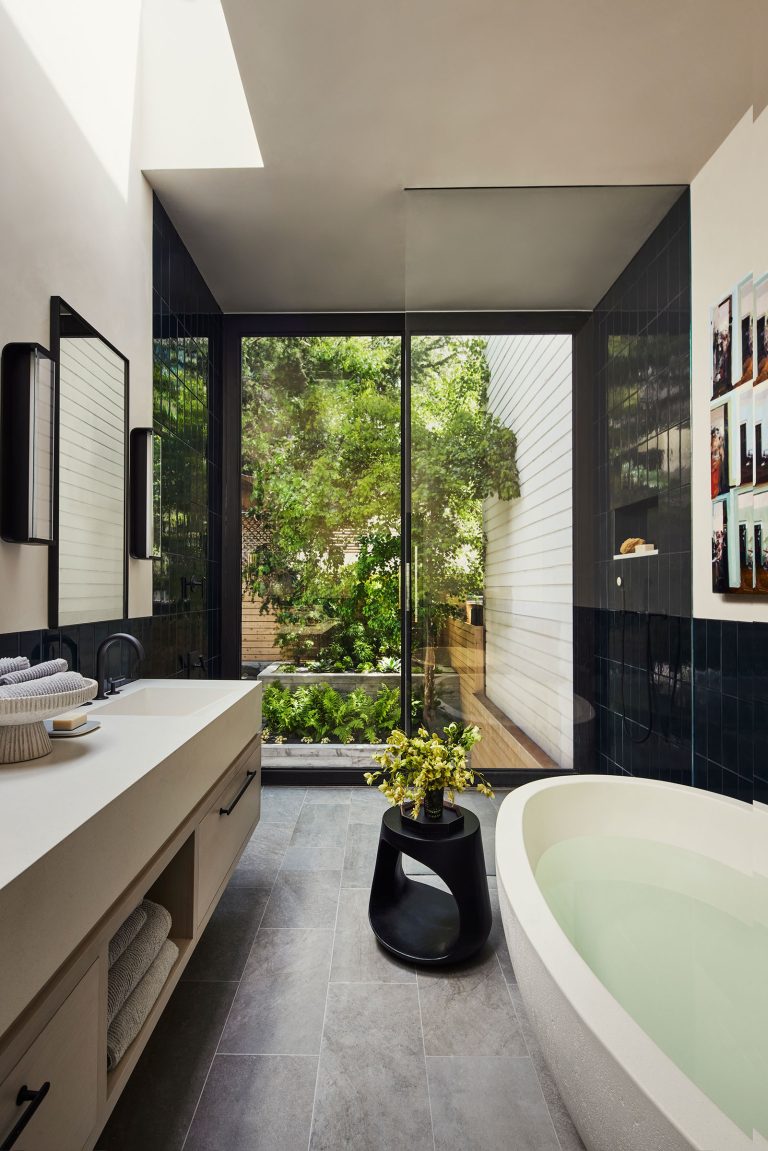 Modern bathroom with gray tile floor, white soaking tub, light vanity with stacked towels, black fixtures, and a small black side table holding a vase of yellow flowers. Large glass doors reveal a lush, green outdoor garden, filling the room with natural light.