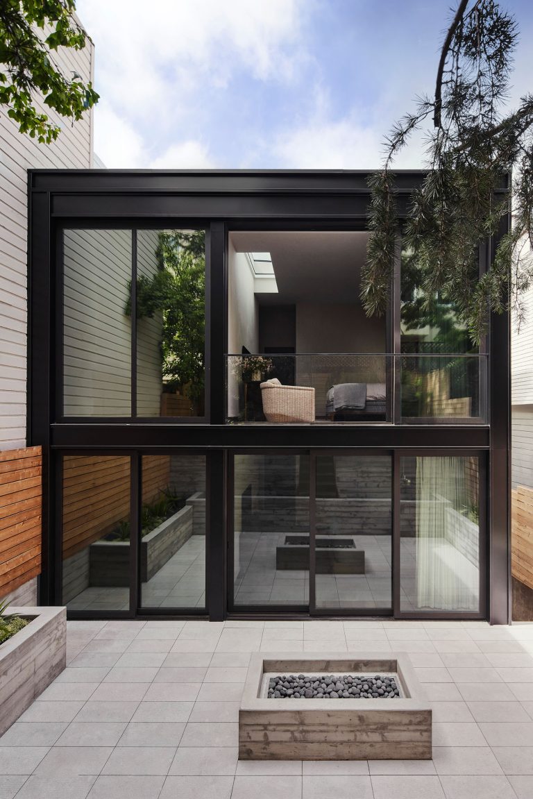 A modern two-story house with large floor-to-ceiling glass windows faces a paved patio with a rectangular fire pit. The house has sleek black frames, minimalist decor, outdoor seating upstairs, and is surrounded by wood fencing and greenery.