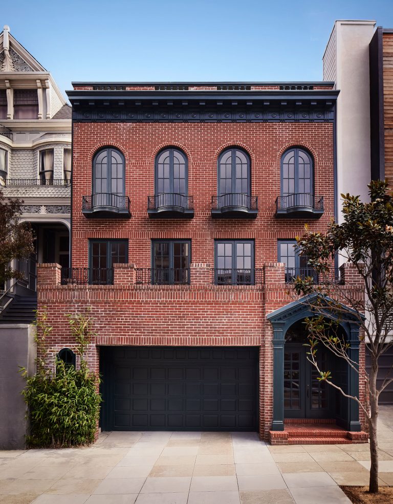 A red brick townhouse with black framed arched windows, black balcony railings, and a large black garage door. There’s a green plant to the left, a small staircase with a dark door on the right, and neighboring houses on both sides.