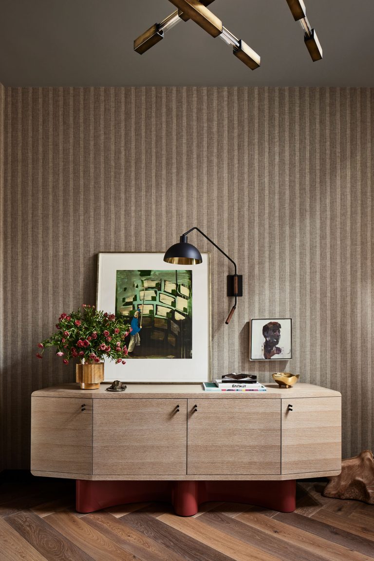 A modern wood sideboard with red legs stands against a striped beige wallpaper. On top are a gold vase with pink flowers, framed art, a small sculpture, books, and a bowl. A black wall lamp hangs above, and part of a geometric ceiling light is visible.