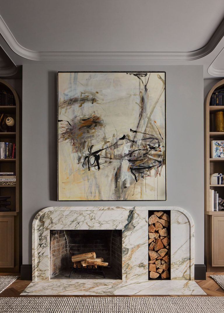 A modern living room features a marble fireplace with stacked firewood to the right. Above the mantel is a large abstract painting in muted tones. Built-in shelves with books and decor flank both sides, and a textured rug covers the floor.