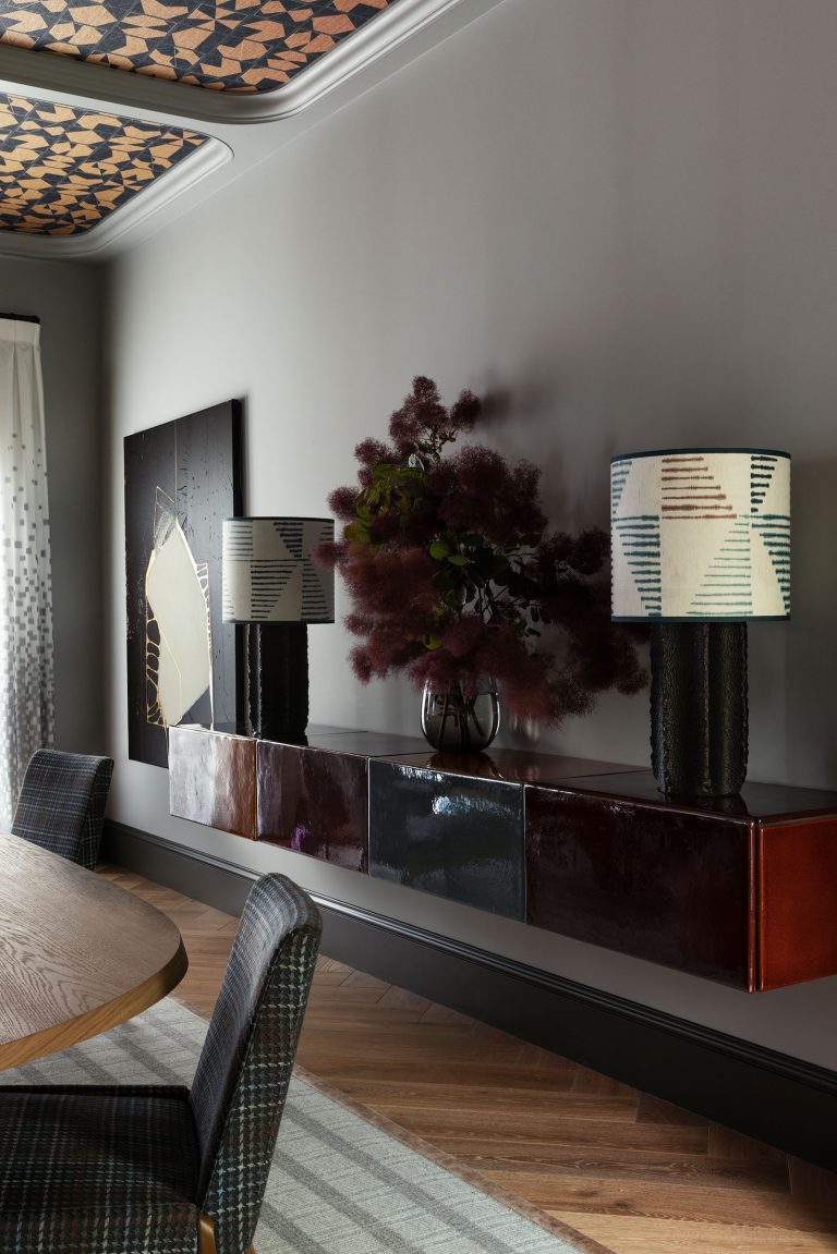 A modern dining room features a floating glossy dark wood sideboard, abstract art, a vase with fluffy dark foliage, two textured black lamps with geometric patterned shades, upholstered chairs, and a wooden table on a striped rug. Ceiling panels have geometric designs.