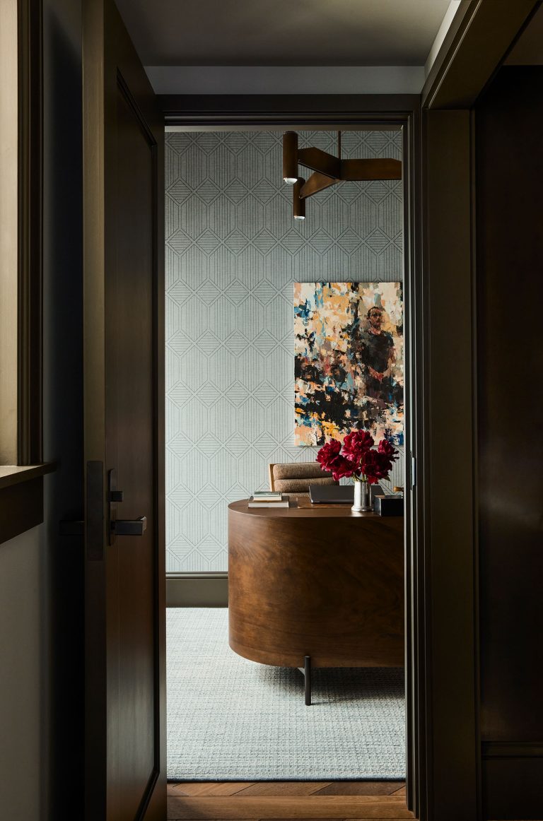 A view through a doorway into a stylish, modern office with a curved wooden desk, a tan chair, a vase of red flowers, geometric wallpaper, abstract wall art, and a contemporary brass light fixture overhead. Natural light softly illuminates the room.