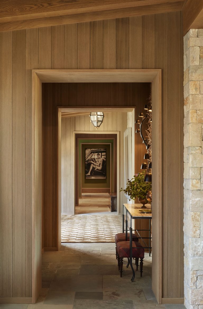 A hallway with wood-paneled walls and stone tile flooring leads through multiple doorways, creating a visual tunnel. A console table with a plant and decorative items stands on the right, and framed artwork hangs on the far wall in the background.