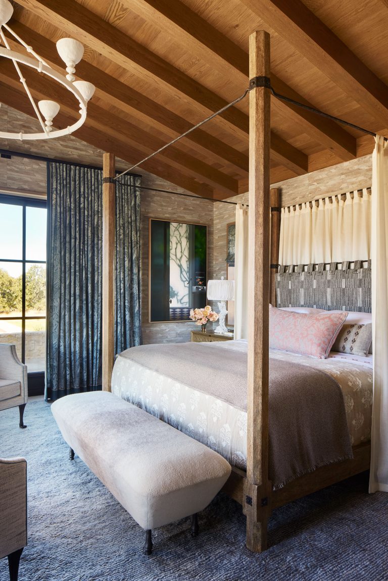A cozy bedroom with a rustic wooden four-poster bed, beige and pastel bedding, a cushioned bench at the foot, large windows with blue curtains, soft lighting, a gray rug, wood-beamed ceiling, armchair, and view of greenery outside.