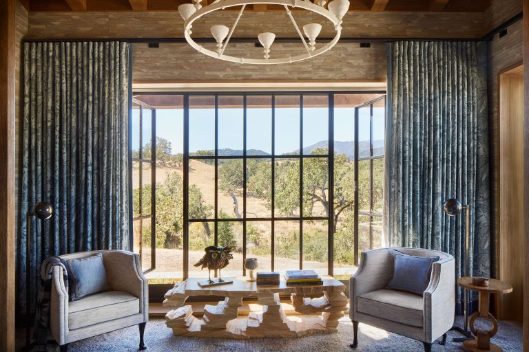 A sunlit room with large windows showing a scenic landscape of hills and trees. Two armchairs face each other across a modern wooden coffee table with books and decor. Blue curtains frame the windows, and a round chandelier hangs from a wood beam ceiling.