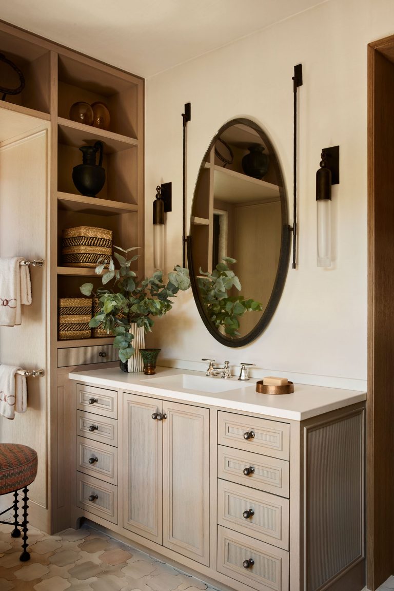 A stylish bathroom vanity with light wood cabinets, an oval mirror, and wall-mounted sconces. A white sink, silver faucet, potted greenery, and decor items sit on the counter. Open shelves on the left display baskets, towels, and pottery. The floor has patterned tiles.