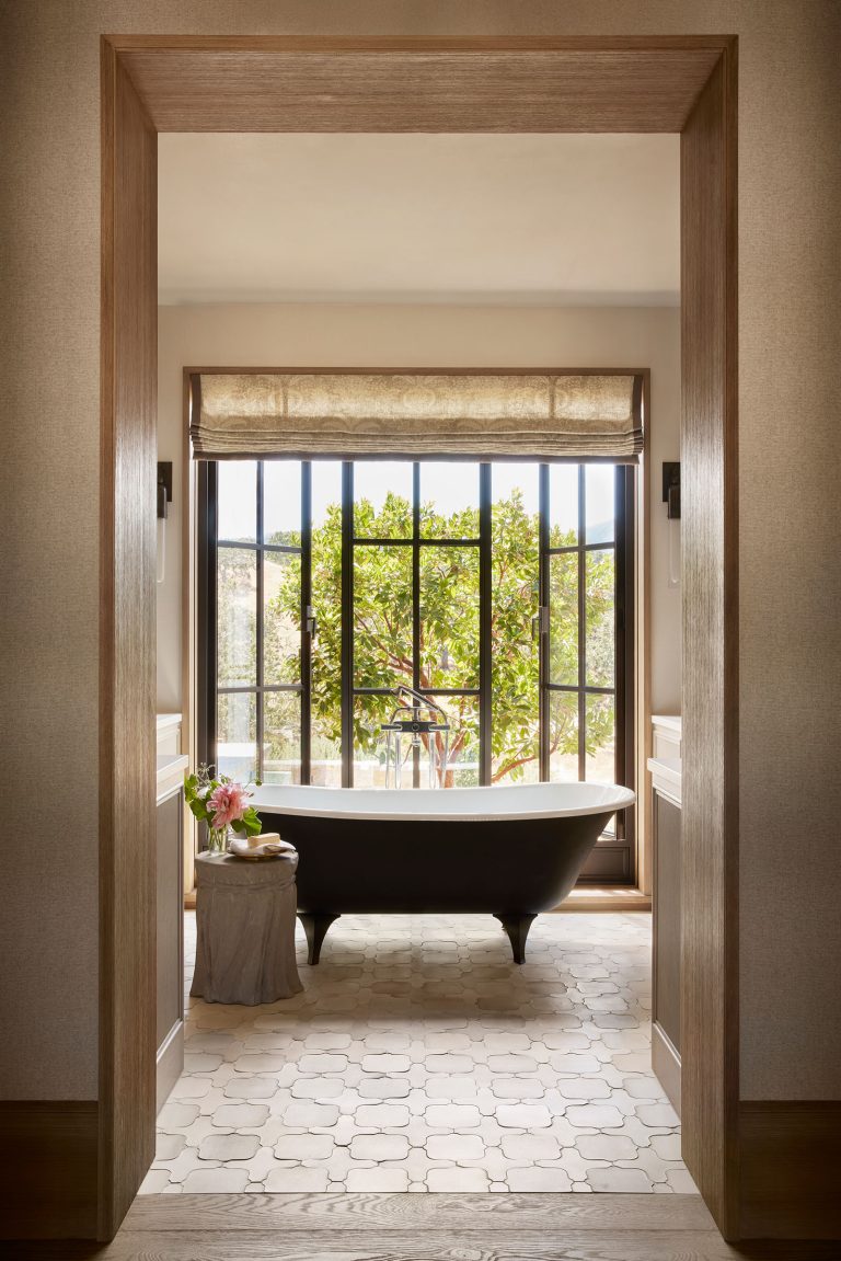 A sunlit bathroom features a black freestanding tub in front of large grid windows with a leafy tree outside. A beige roman shade tops the window. A small table with a vase, pink flower, and towel sits beside the tub on patterned tile flooring.