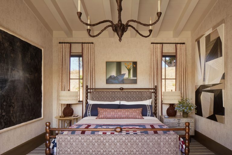 A stylish bedroom with a wooden bed centered under a dramatic dark chandelier. The bed has patterned pillows and a patchwork quilt. Abstract art hangs above the headboard and on surrounding walls. Two nightstands with lamps and a vase of flowers sit beside the bed.