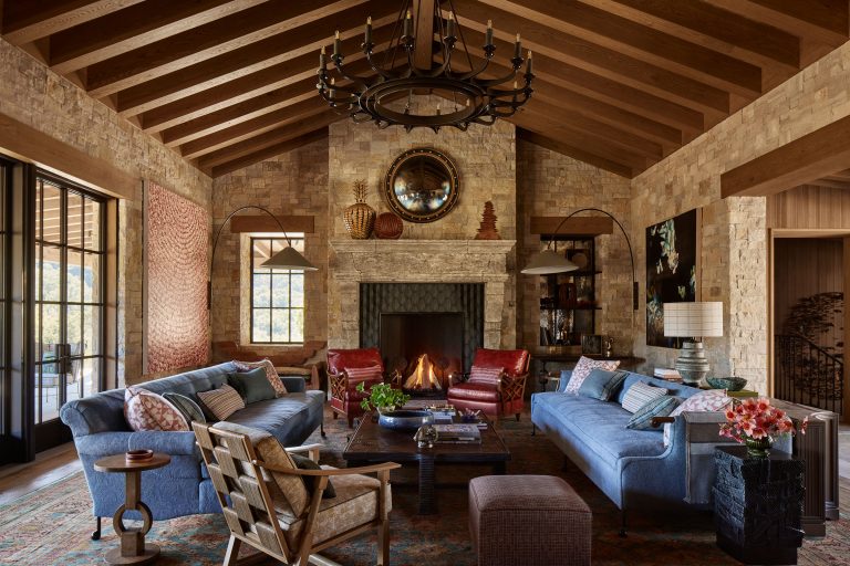 A rustic living room with exposed wooden beams, stone walls, and a lit fireplace. Two blue sofas face each other, flanked by armchairs. A large round mirror hangs above the mantel. There are decorative lamps, artwork, and large windows letting in natural light.