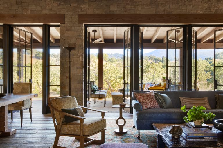 A cozy living room with rustic stone walls, glass doors opening to a sunny patio, and views of trees and hills. The room features a wooden armchair, a blue sofa with patterned pillows, a coffee table, and warm, natural light filling the space.