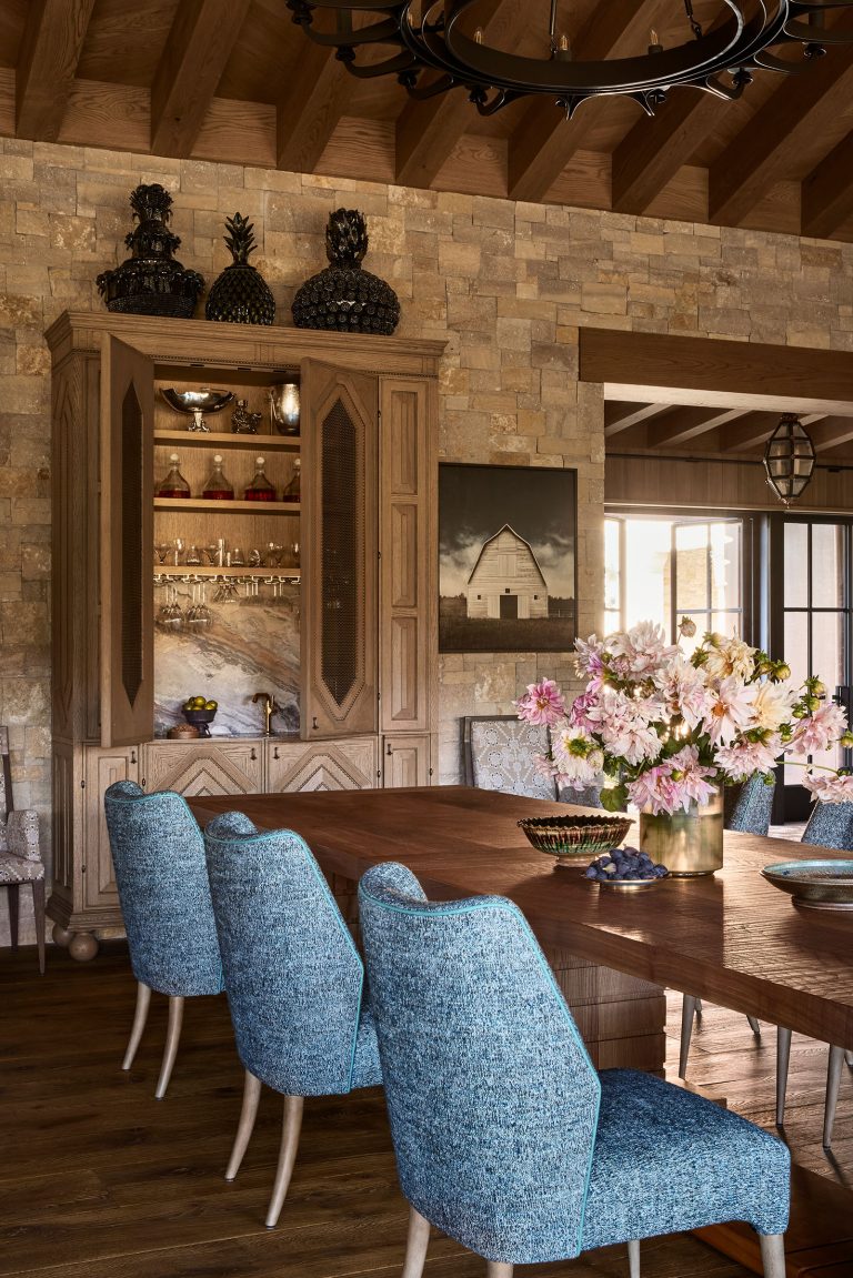 A rustic dining room features a wooden table with blue upholstered chairs, a large bouquet of pink flowers, a stone wall, a wooden cabinet with glassware, decorative busts on top, and a barn photograph hanging on the wall. Warm, natural light fills the space.