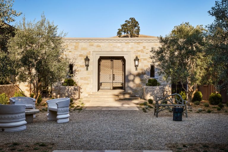 A stone building has a central arched doorway with double doors and two large lanterns. The courtyard features gravel, trimmed bushes, olive trees, white sculpted outdoor chairs, a curved garden bench, and a small round table.