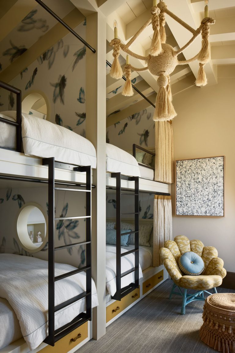 A cozy bedroom with two sets of white bunk beds, black ladders, round windows, and under-bed drawers. The wallpaper features blue-green feathers. Theres a unique tassel chandelier and a yellow armchair with a blue floral cushion. A textured wall art hangs nearby.